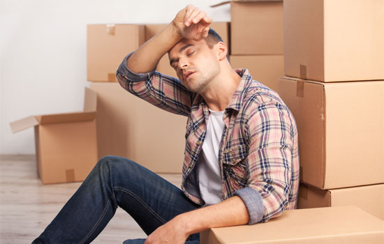 Man sitting against moving boxes wiping his sweaty forehead